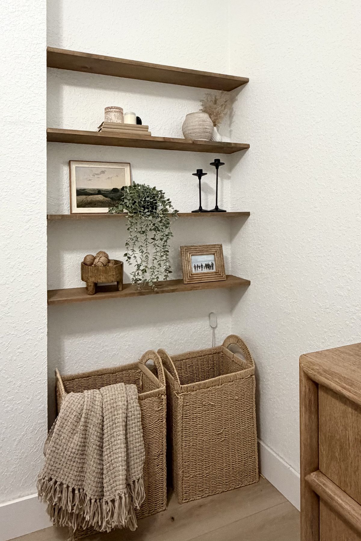 The floating shelf display in a wall nook. 4 wooden shelves, installed and decorated with plants, images, candle sticks, and vases.